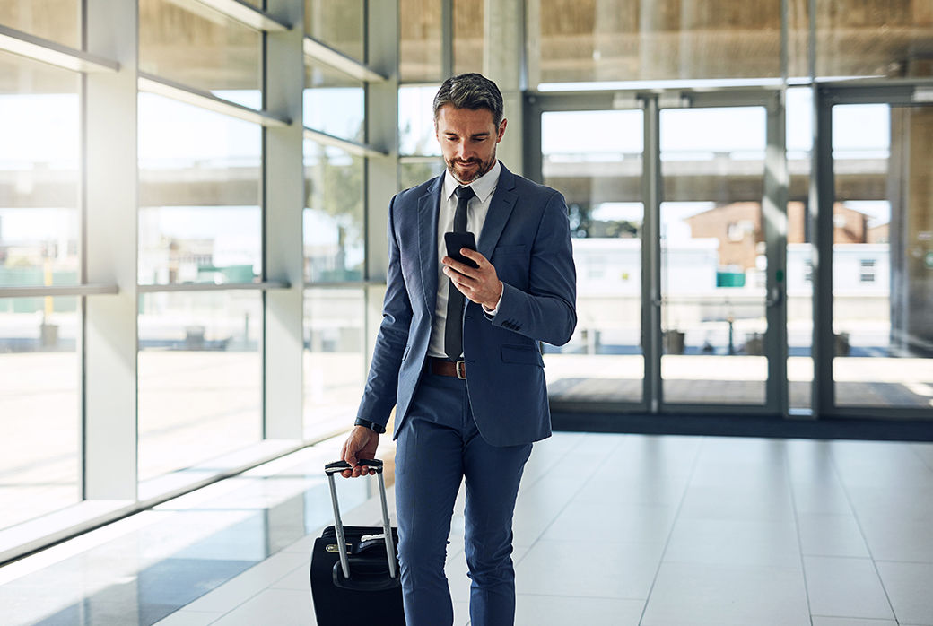 Man in a suit checking his phone while pulling a suitcase through an airport